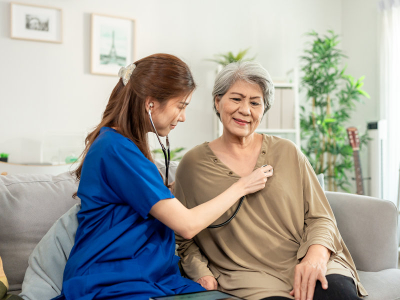 Healthcare worker in blue scrubs using a stethoscope to check the chest of an elderly woman sitting on a couch at home.