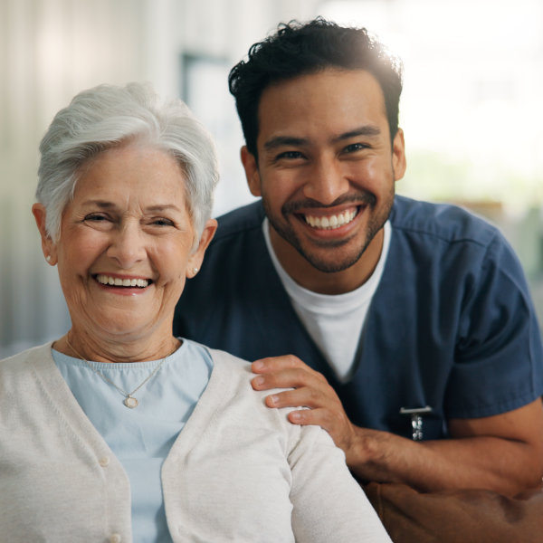 Smiling elderly woman with short white hair sitting next to a cheerful male nurse in scrubs with his hand on her shoulder.