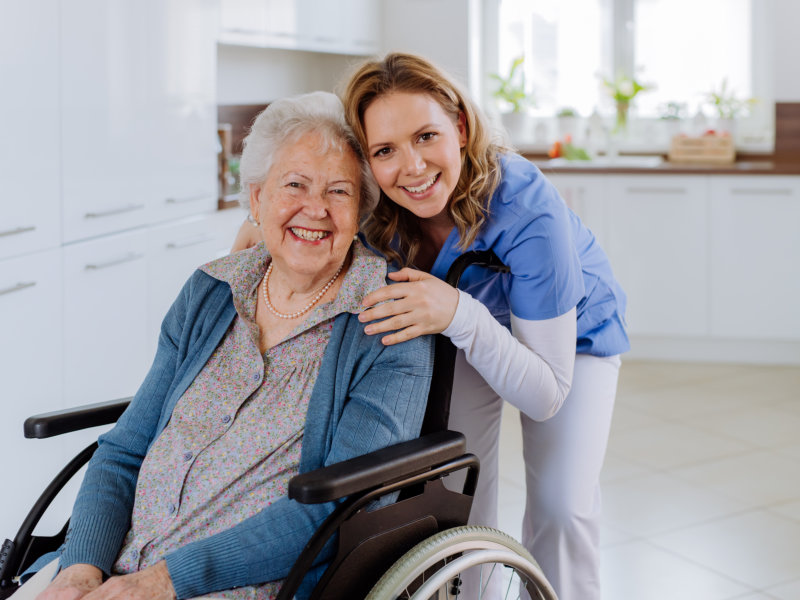 Smiling elderly woman in a wheelchair with a female healthcare worker embracing her in a bright, modern kitchen.