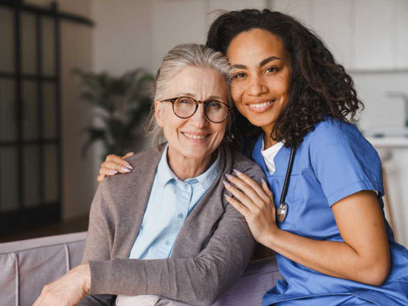 Smiling nurse in blue scrubs embraces elderly woman with glasses, both seated and looking warmly at the camera.