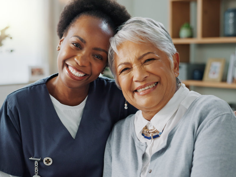 Smiling nurse and elderly woman sitting closely, showing warmth and companionship in a cozy indoor setting
