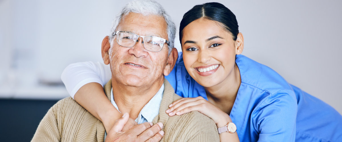 Elderly man with glasses smiling, embraced from behind by a smiling young woman in blue scrubs, showing care and warmth.