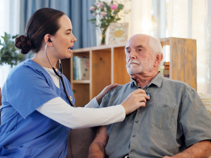 Healthcare worker in blue scrubs uses a stethoscope to check the heartbeat of an elderly man sitting on a couch at home.