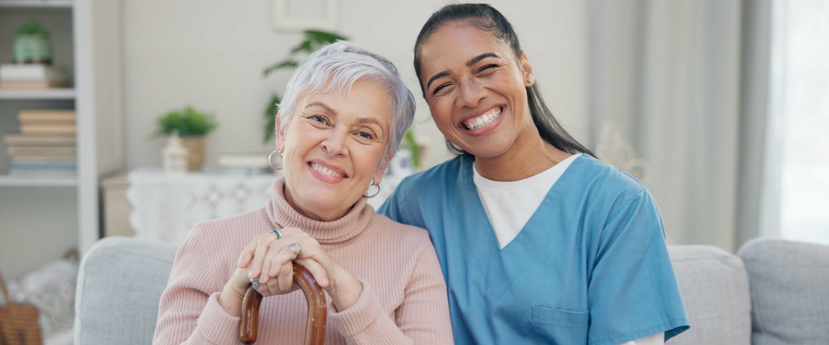 Smiling elderly woman with a cane sitting next to a cheerful caregiver in blue scrubs on a cozy couch indoors.