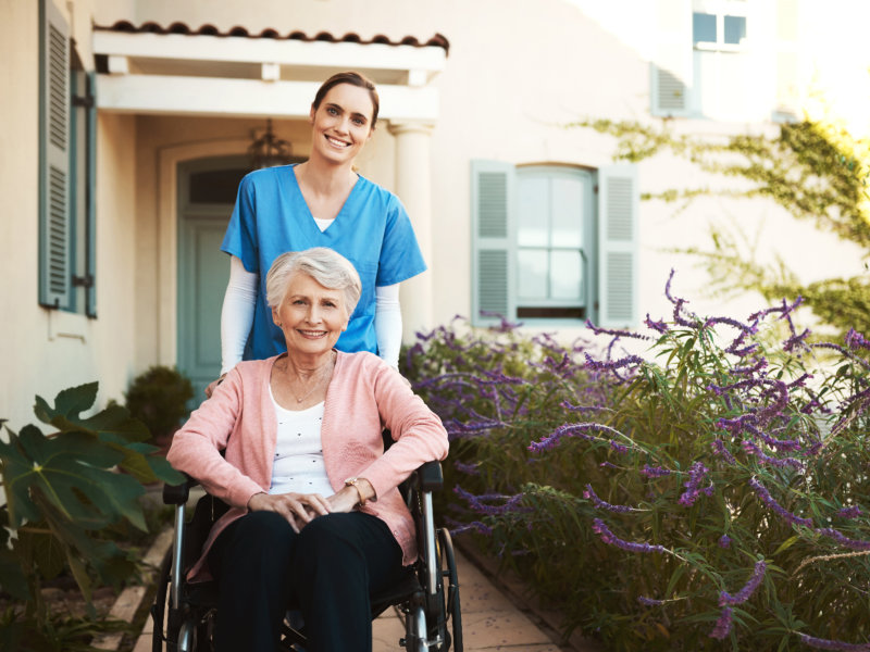Caregiver in blue scrubs smiles behind elderly woman in wheelchair outside a house with purple flowers and greenery.