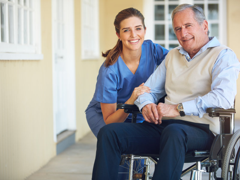 Young female caregiver smiling beside an elderly man in a wheelchair outside a building