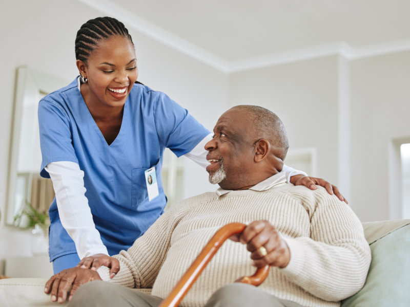 A smiling nurse in blue scrubs gently holds the hand of an elderly man with a cane, both sharing a warm moment indoors.