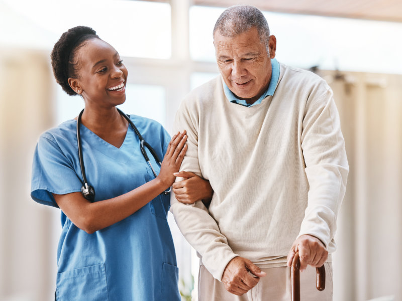 A smiling Black female healthcare worker in blue scrubs, wearing a stethoscope around her neck, gently supports an older man by the arm as he walks using a wooden cane.