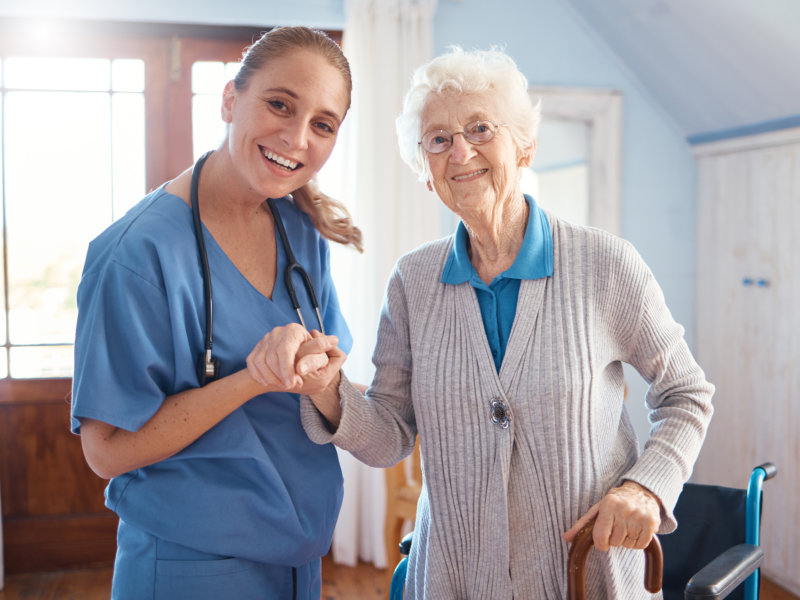 A smiling female healthcare worker in blue scrubs, wearing a stethoscope around her neck, stands holding the hand of an elderly woman who is supported by a cane and positioned near a wheelchair.