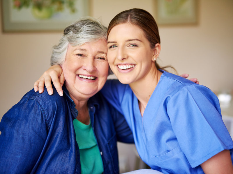 Smiling elderly woman and healthcare worker in blue scrubs hugging and posing closely together indoors.