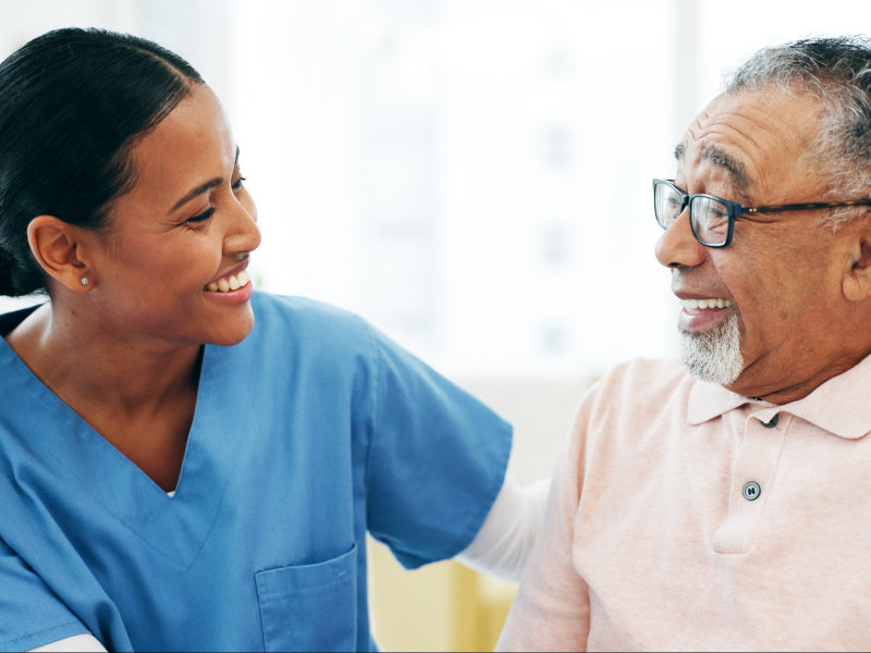 A smiling nurse in blue scrubs warmly interacts with an elderly man wearing glasses and a light pink polo shirt.