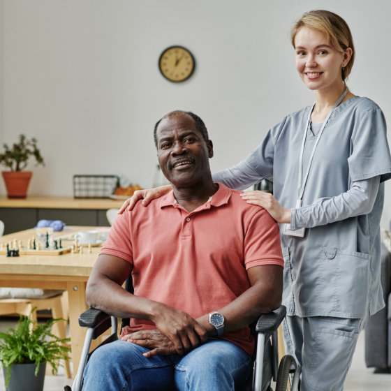 Patient in a wheelchair smiling with a nurse standing beside him, hand on his shoulder, in a bright room with a clock and plants.