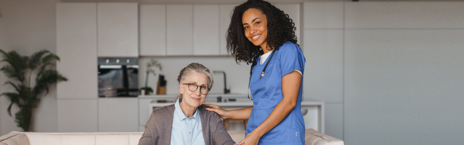A smiling caregiver with a stethoscope rests a supportive hand on the shoulder of an elderly woman who is sitting on a sofa.