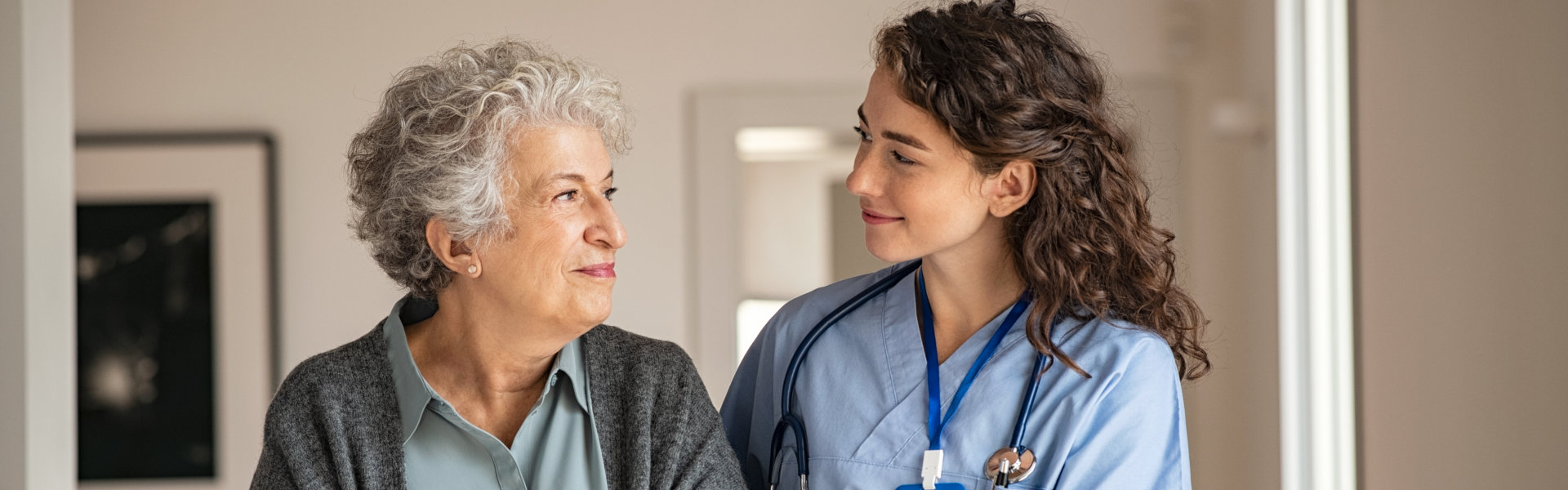 A caring nurse with a stethoscope looks toward an elderly woman, who smiles gently in a supportive, professional environment.