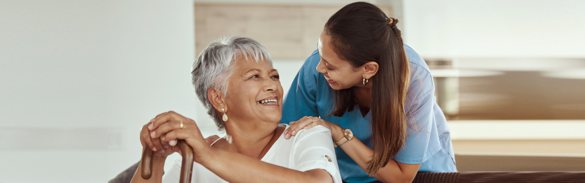 A smiling caregiver rests a hand on the shoulder of an elderly woman, who holds a walking cane and looks back at her with a joyful expression.
