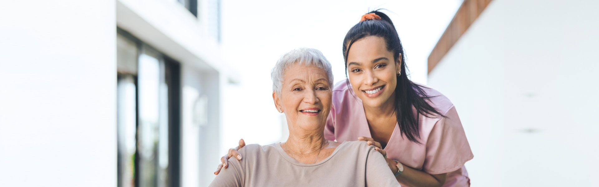 Smiling elderly woman with short gray hair and younger woman in pink scrubs with ponytail outdoors, showing support and care.